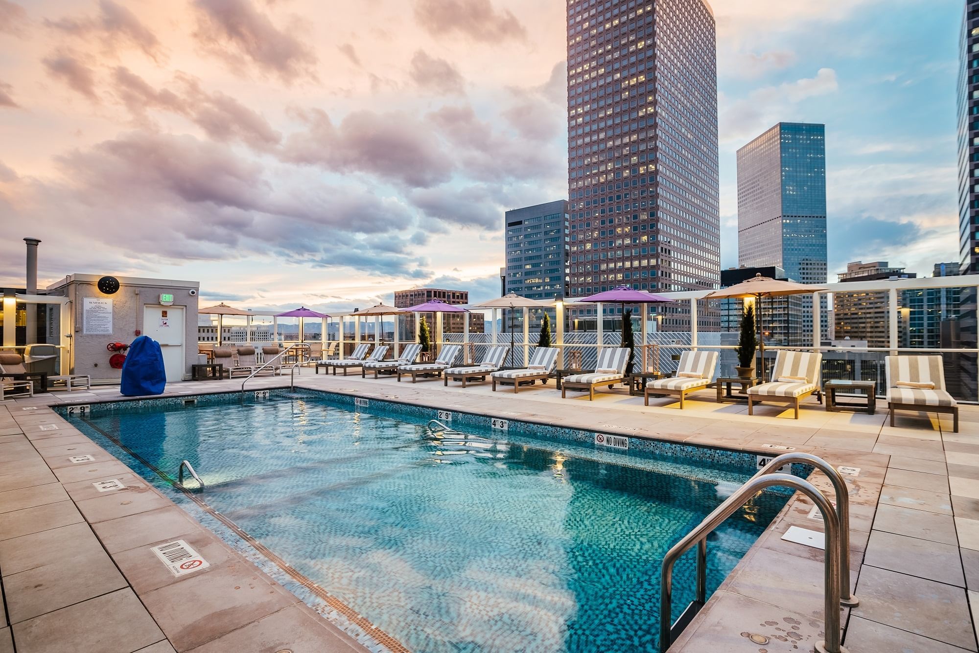 A rooftop pool with lounge chairs and umbrellas overlooking the city skyline at sunset.