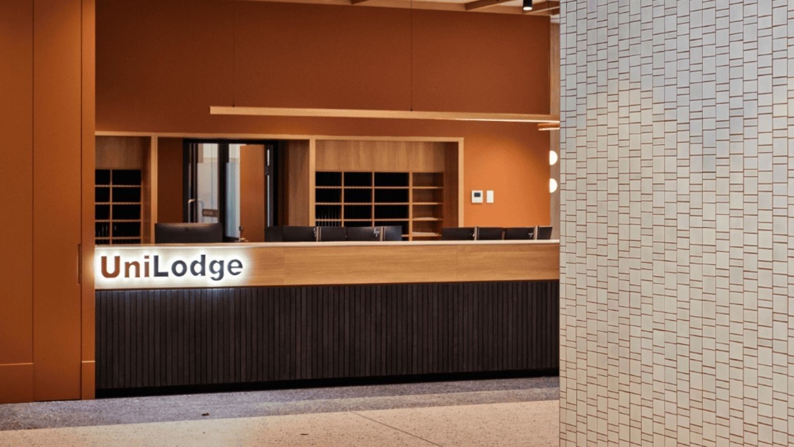 UniLodge reception area with wooden counter and black chairs, orange walls, and tiled column.