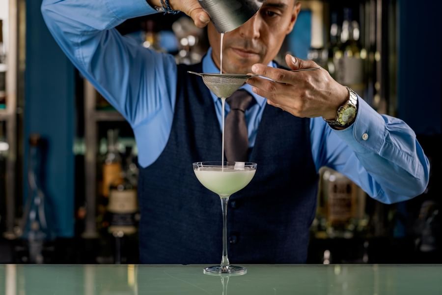 Bartender straining green cocktail through sieve into stemware over counter at Tom Brown at The Capital