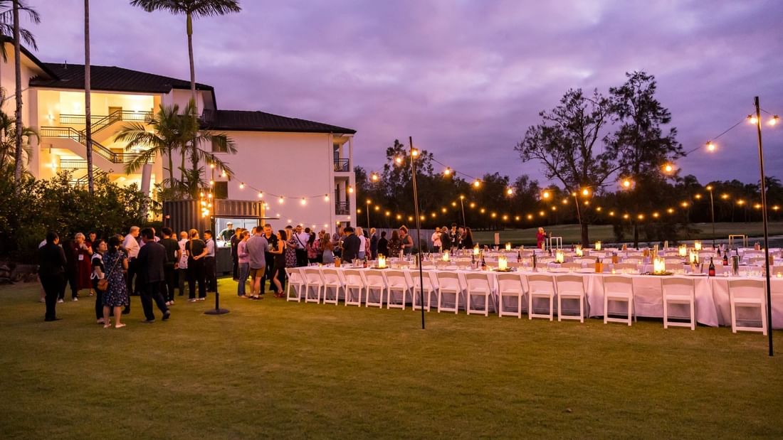 A large outdoor event with tables and chairs set up on a lawn, surrounded by string lights and palm trees.