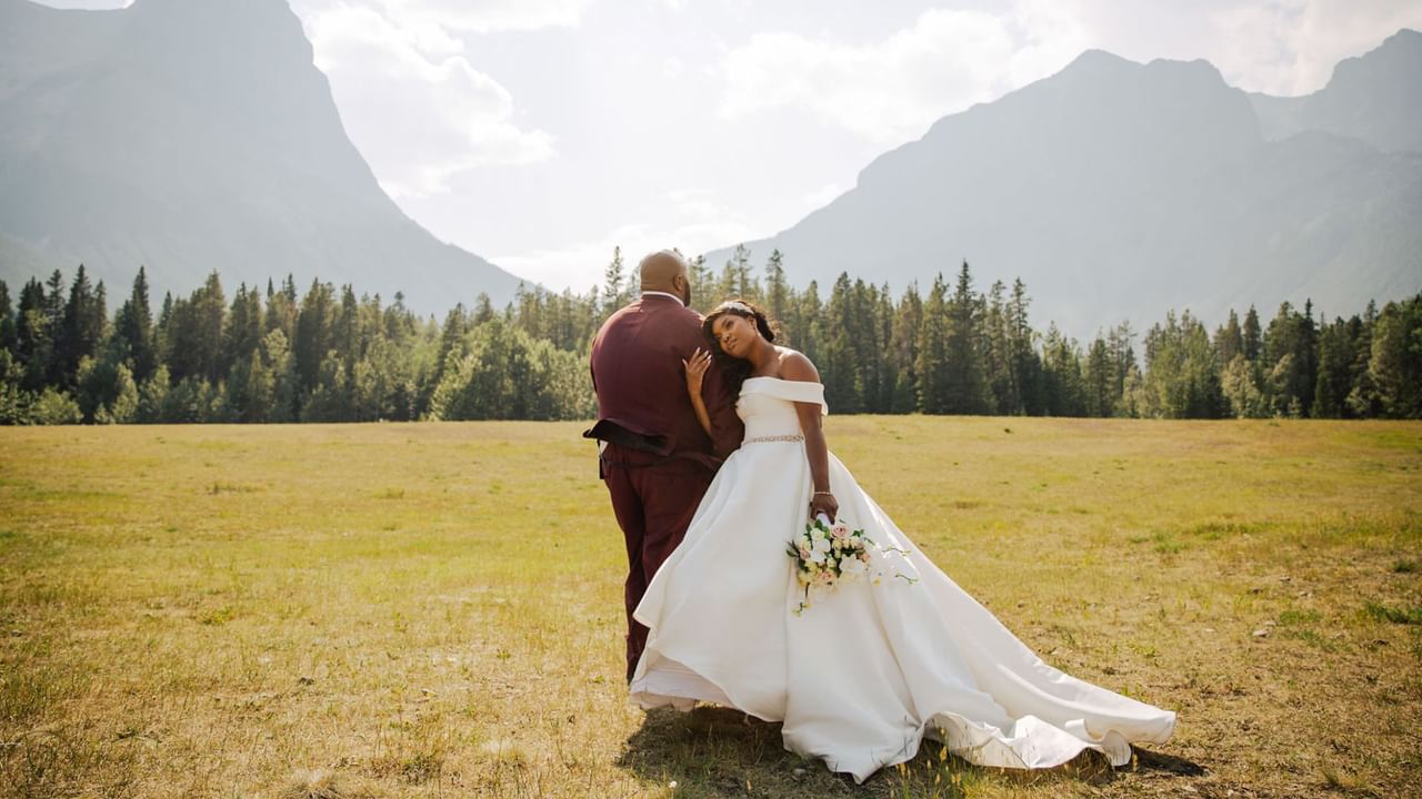 A bride and groom standing in a meadow with mountains and trees in the background.