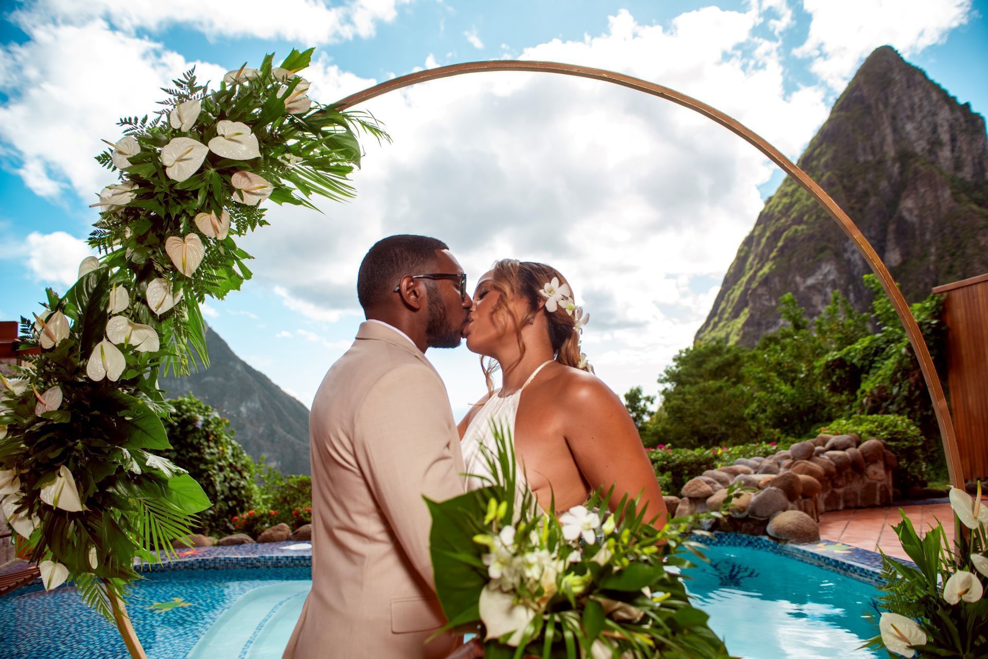 A couple kisses under a floral arch by a pool with mountains in the background.