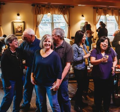 A group of people are gathered in a social setting, laughing and interacting while holding drinks.