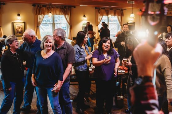 A group of people are gathered in a social setting, laughing and interacting while holding drinks.