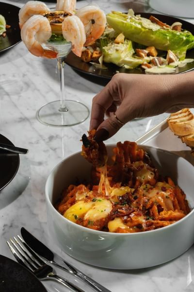 Hand dipping into a bowl of cheesy Frankfort Poutine, served with shrimp cocktail and fresh salad at Cypress & Oak
