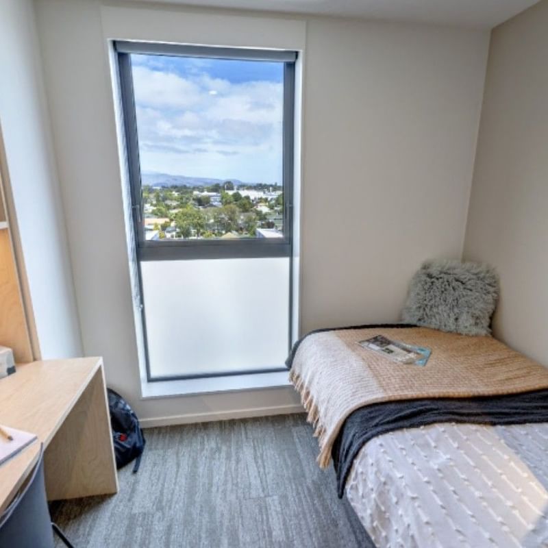 Compact guest room with single bed, built-in desk, shelving, and window overlooking suburban landscape at Tupuārangi