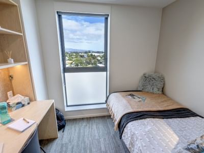 Compact guest room with single bed, built-in desk, shelving, and window overlooking suburban landscape at Tupuārangi
