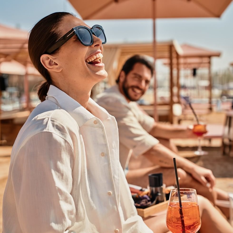 Coppia sorridente che gode di un drink in spiaggia con ombrellone, promo Prebook & Save