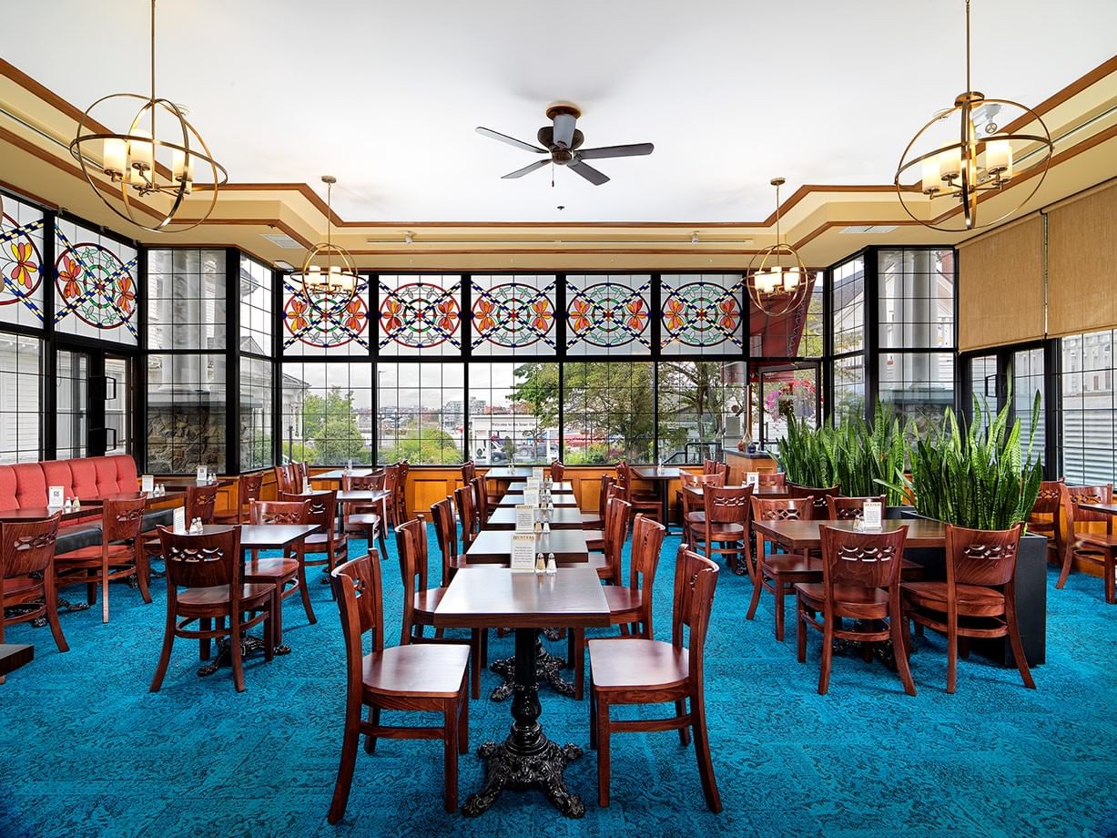 A dining room with blue carpet, multiple tables, wooden chairs, stained glass windows, and large globe-shaped ceiling lights.