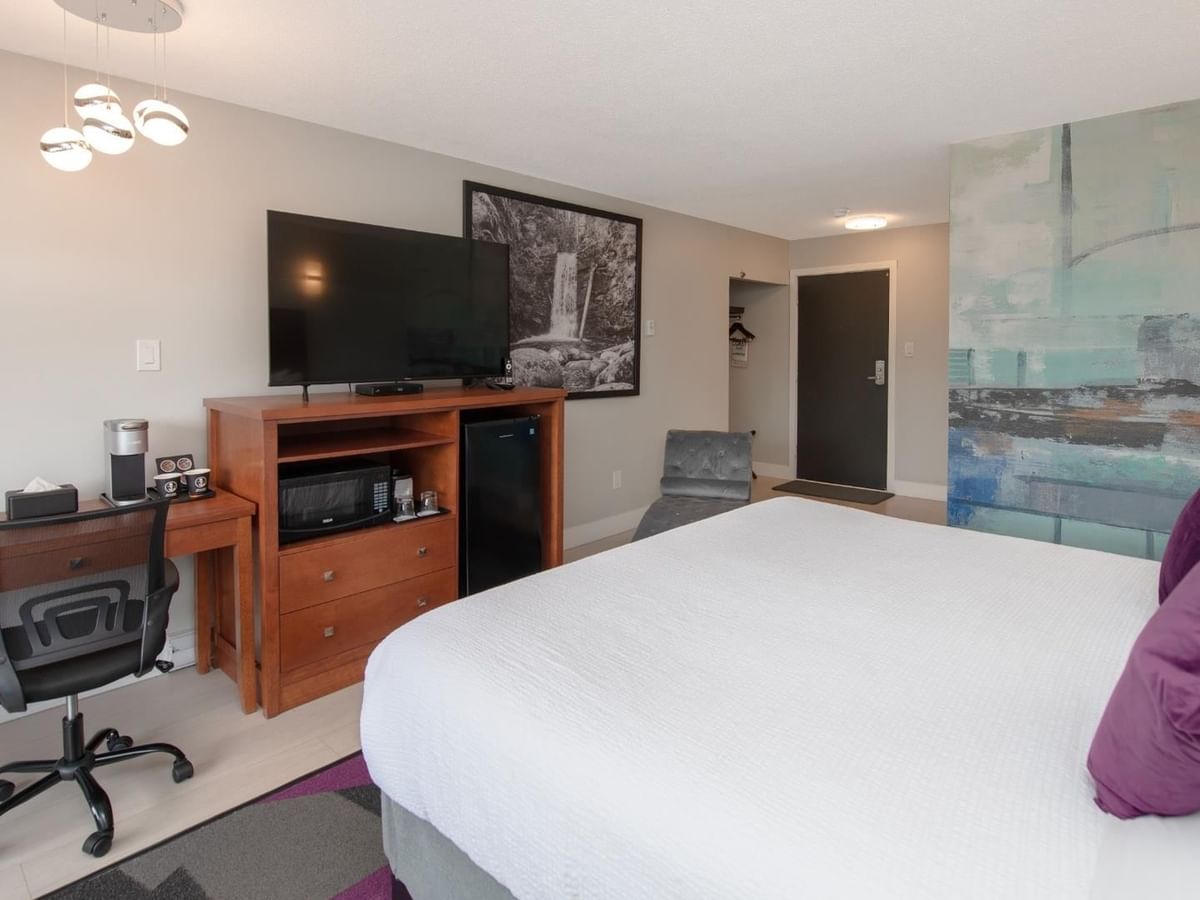 Hotel room with large bed, purple pillows, desk, chair, TV, and colorful wall art.