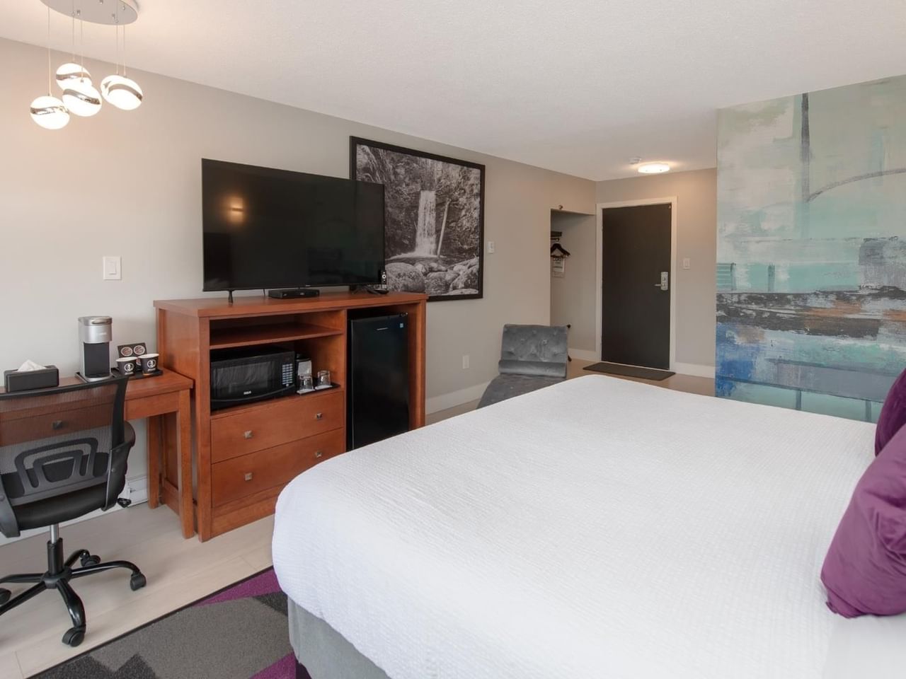 Modern hotel king room with a bed, desk, TV, and abstract art on the wall.