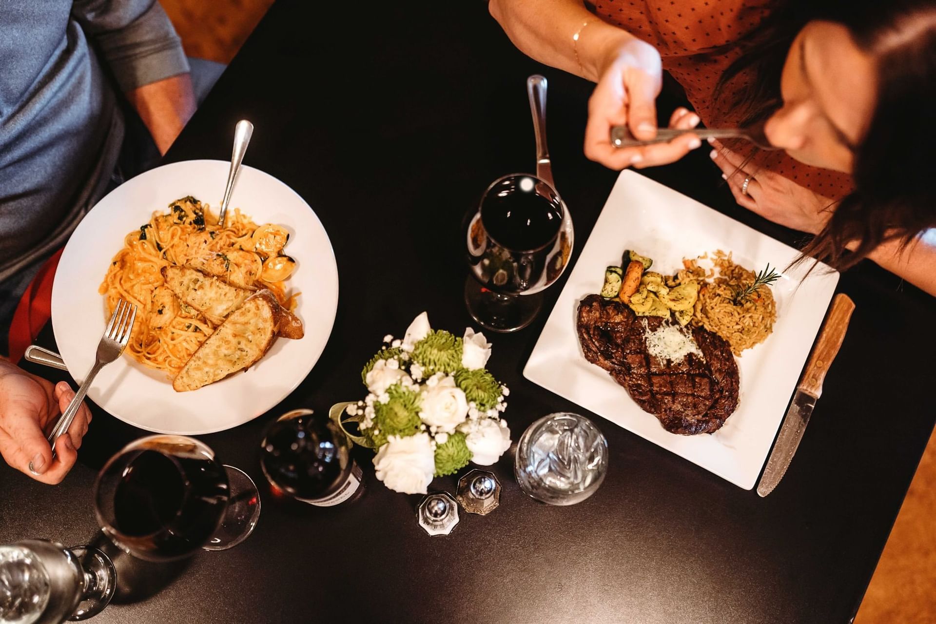 Two people dining with plates of pasta, steak, wine, and a floral centerpiece on a dark table.