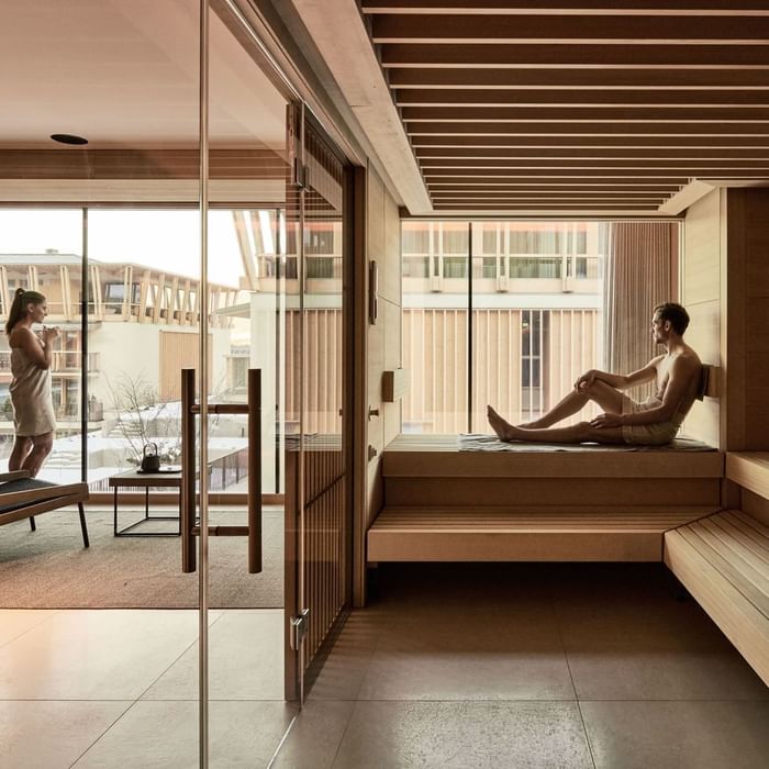 Mann sitzt in Sauna mit Glaswänden, Frau steht draußen, Gebäude und Bäume im Hintergrund.