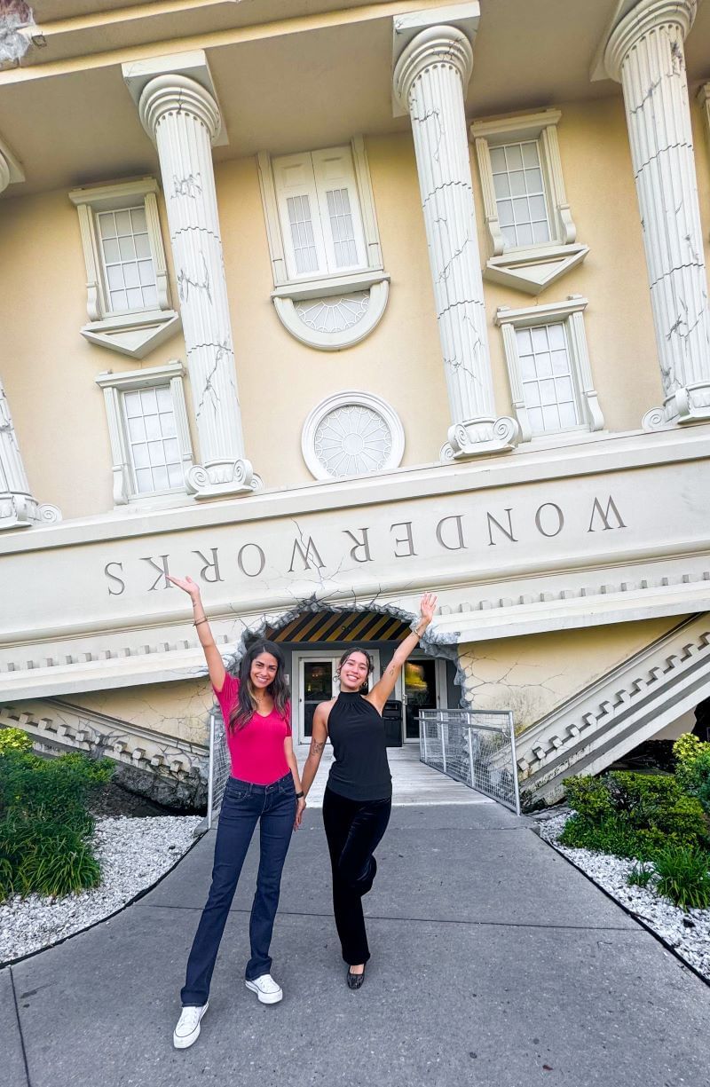 Two women smiling and posing in front of an upside down light yellow building called WONDERWORKS. 