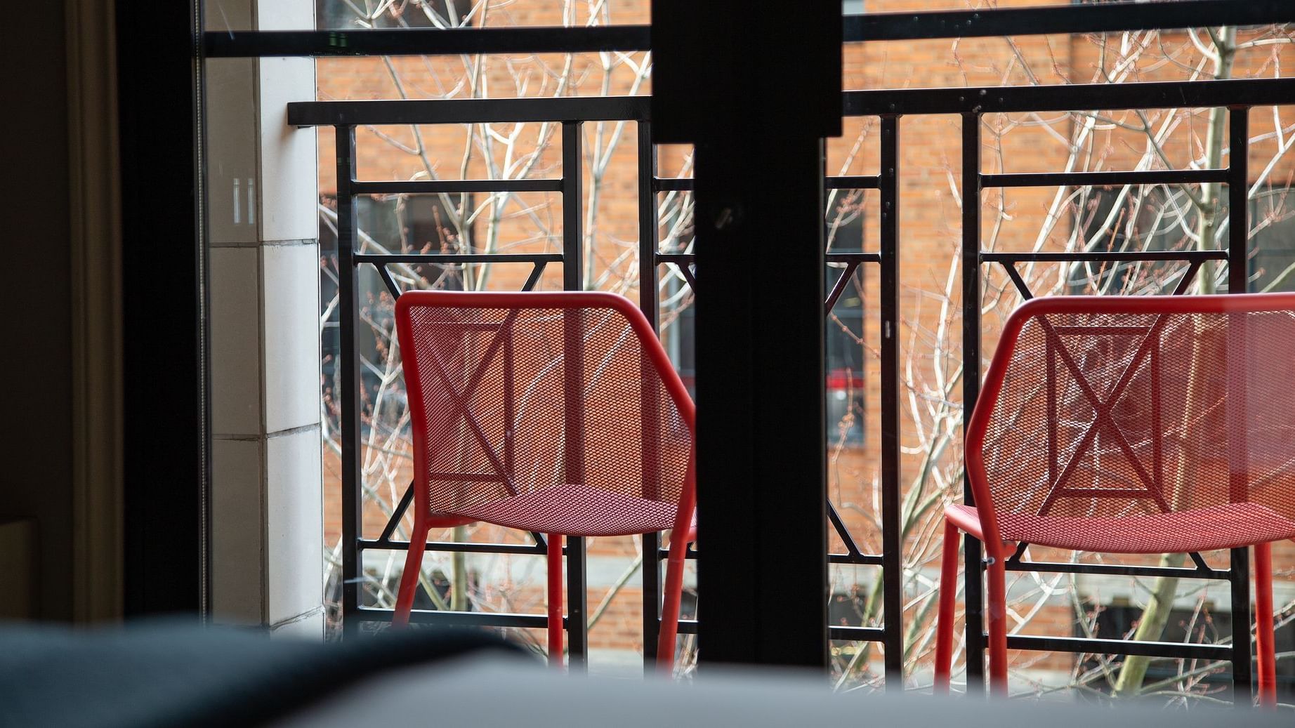 Two red chairs sit on a balcony with a black railing, overlooking a brick building and bare trees.