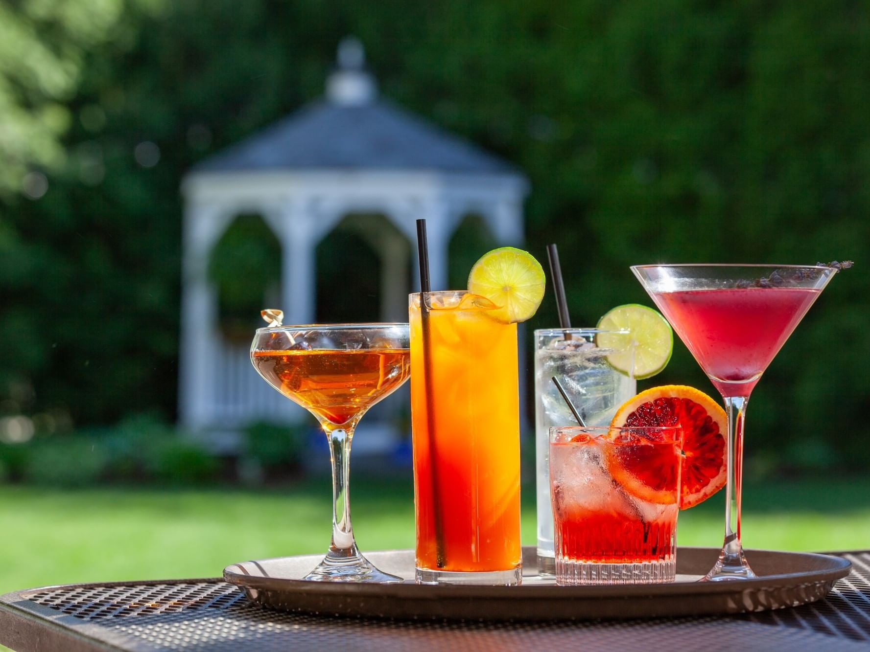 Four colorful cocktails with citrus garnishes displayed on a wooden tray in a lush garden setting with a gazebo in the background.