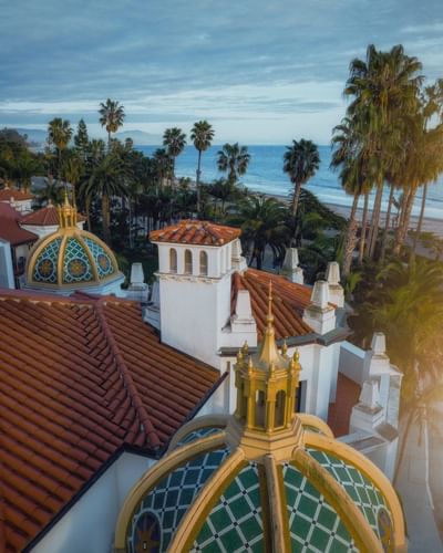 Aerial view of Santa Barbara Inn’s dome rooftops, nestled among palm trees, overlooking a calm ocean under a cloudy sky