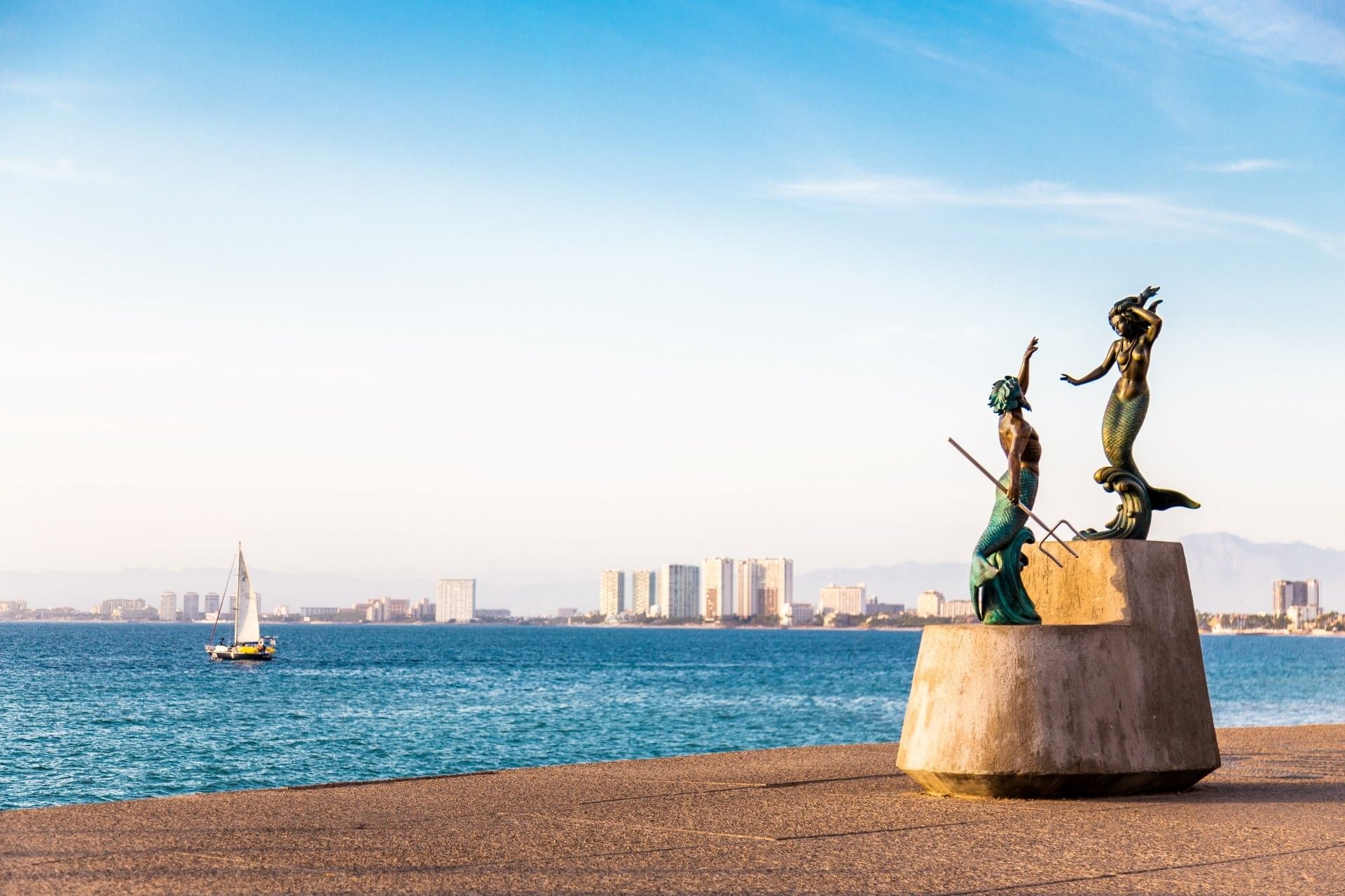 The Malecón in Puerto Vallarta featuring the iconic Triton and Mermaid sculpture overlooking the ocean, with a sailboat and city skyline in the background.