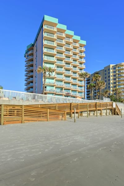 Daytona Bahama House beachfront condo with balcony, wooden ramp, and palm trees under a clear blue sky.