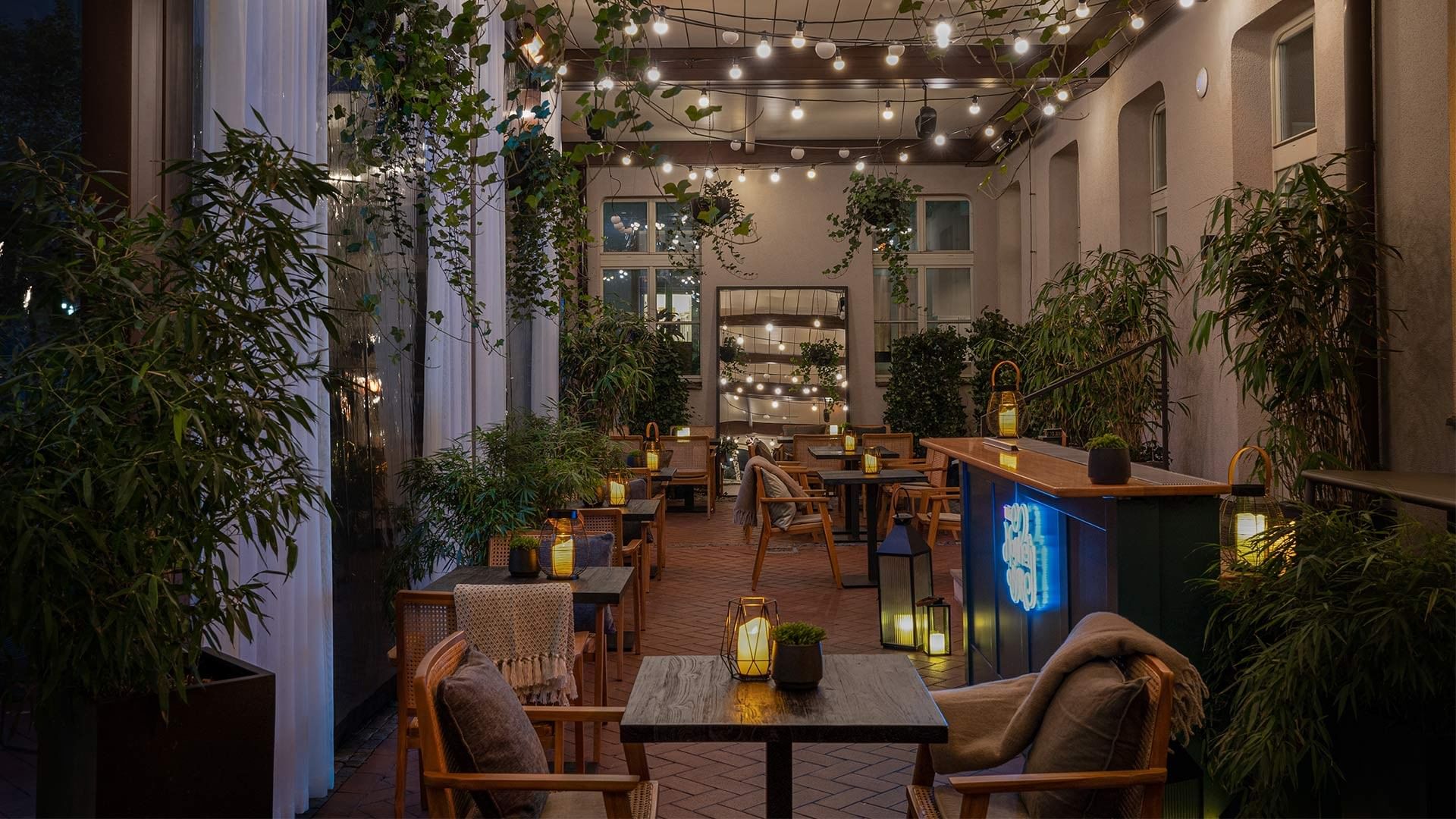 An outdoor dining area with tables, chairs, string lights, and lanterns, surrounded by plants and a bar counter.