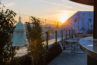 Rooftop terrace with a glass railing, vintage white chairs, and a view of the Mariahilfer-Hof dome at Hotel Motto Vienna
