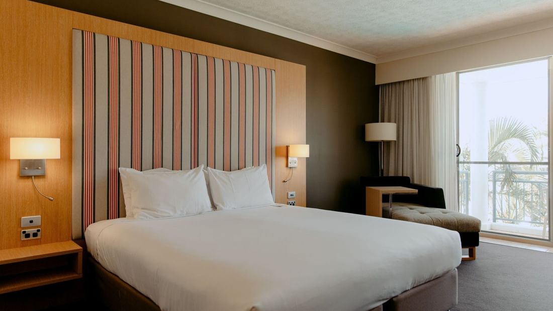 A hotel room with a large bed, striped headboard, two lamps, and a balcony with a view.