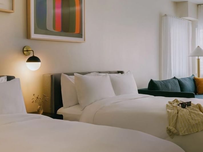 Two beds with white linens, pillows, and a couch with pillows in a room with a wall painting and a lamp.