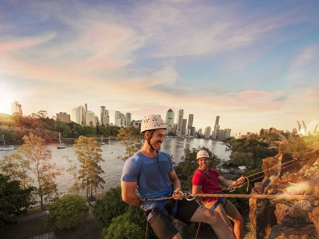 Two men abseiling the Kangaroo Point Cliffs at sunset with the city skyline in the background near The Sebel Brisbane