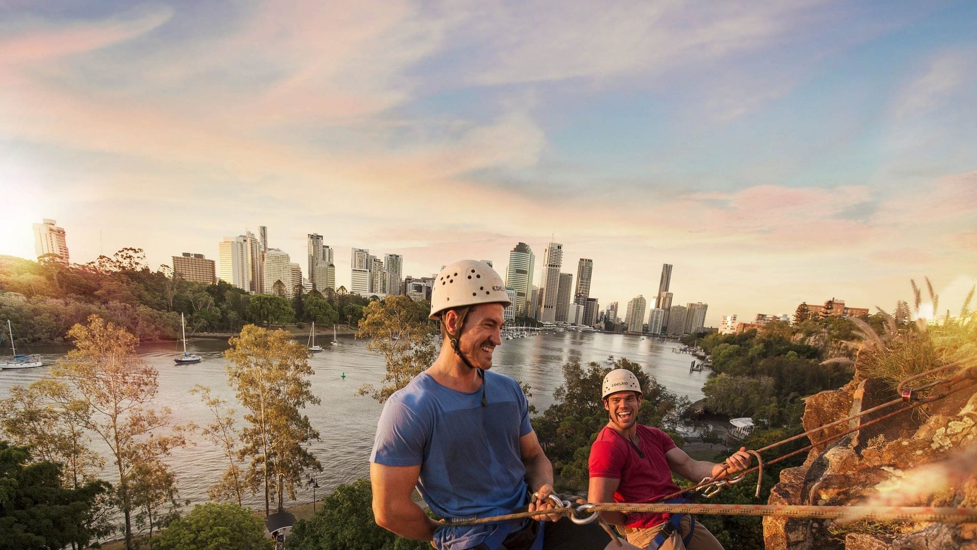 Two men abseiling the Kangaroo Point Cliffs at sunset with the city skyline in the background near The Sebel Brisbane