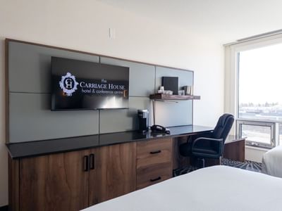 Modern hotel room with desk, chair, flat screen TV, and window at Carriage House Hotel & Conference Centre.