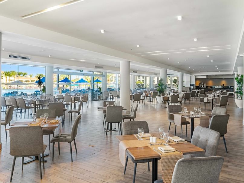 A spacious restaurant with wooden floors, white ceilings, and large windows showing an outdoor pool area.