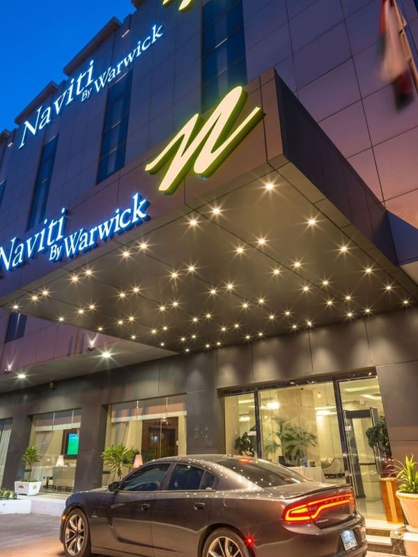 Vue extérieure de l'hôtel Naviti by Warwick Al Khobar, avec une voiture sombre garée sous un auvent lumineux