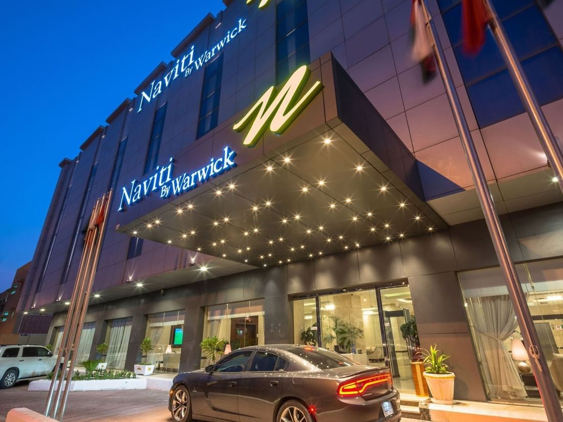 Vue extérieure de l'hôtel Naviti by Warwick Al Khobar, avec une voiture sombre garée sous un auvent lumineux