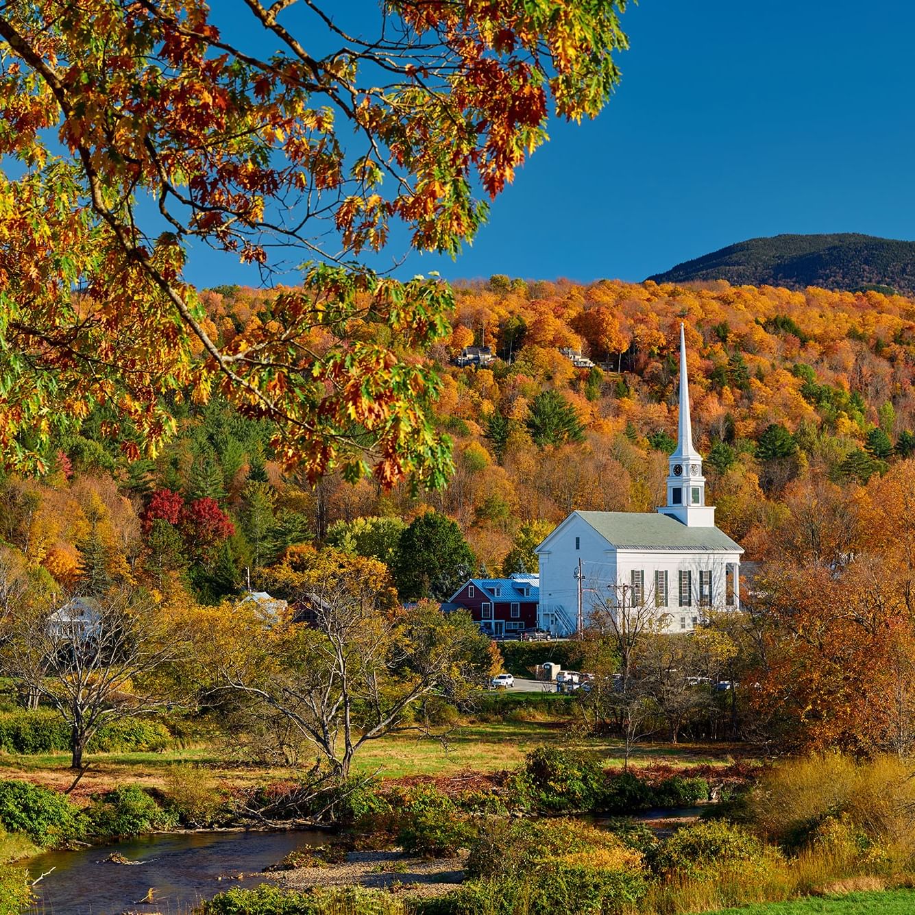 White church with steeple among autumn trees and a small stream under a clear blue sky.