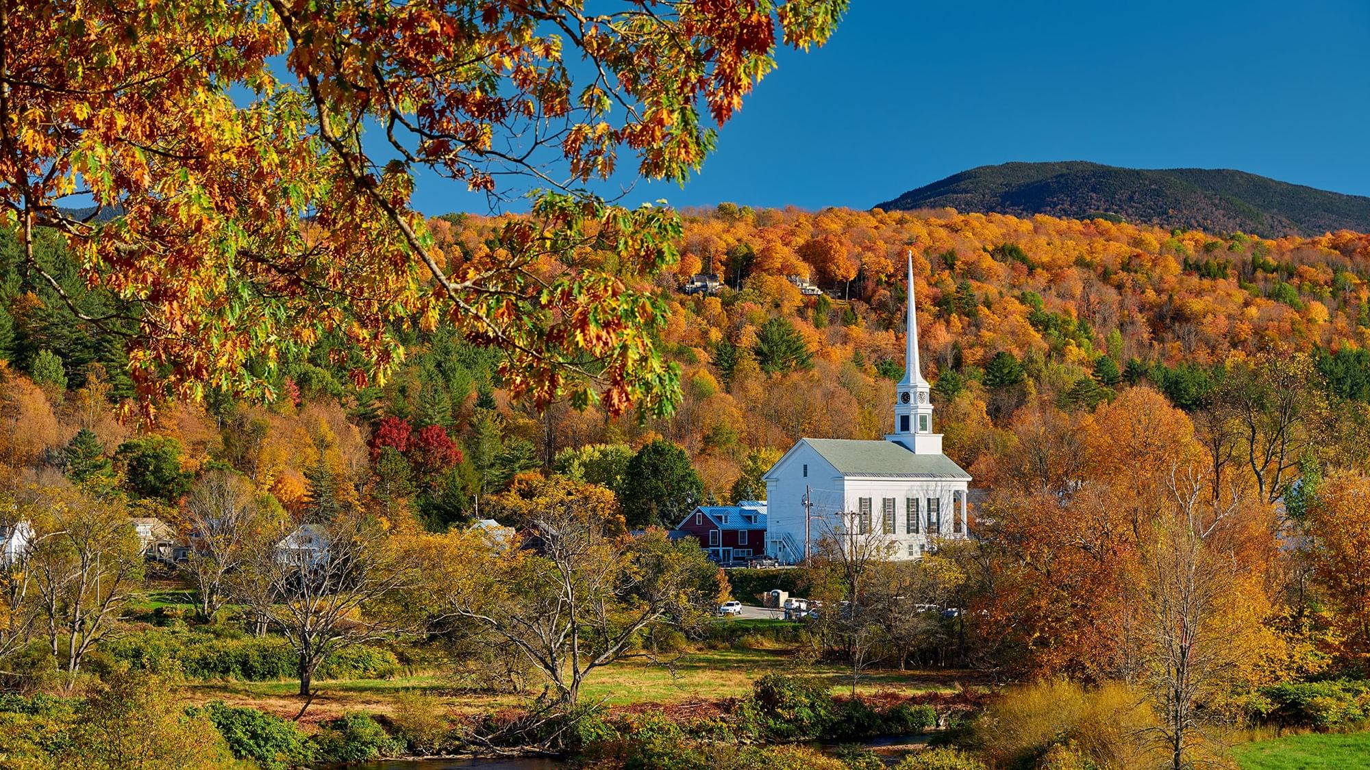 White church with steeple among autumn trees and a small stream under a clear blue sky.