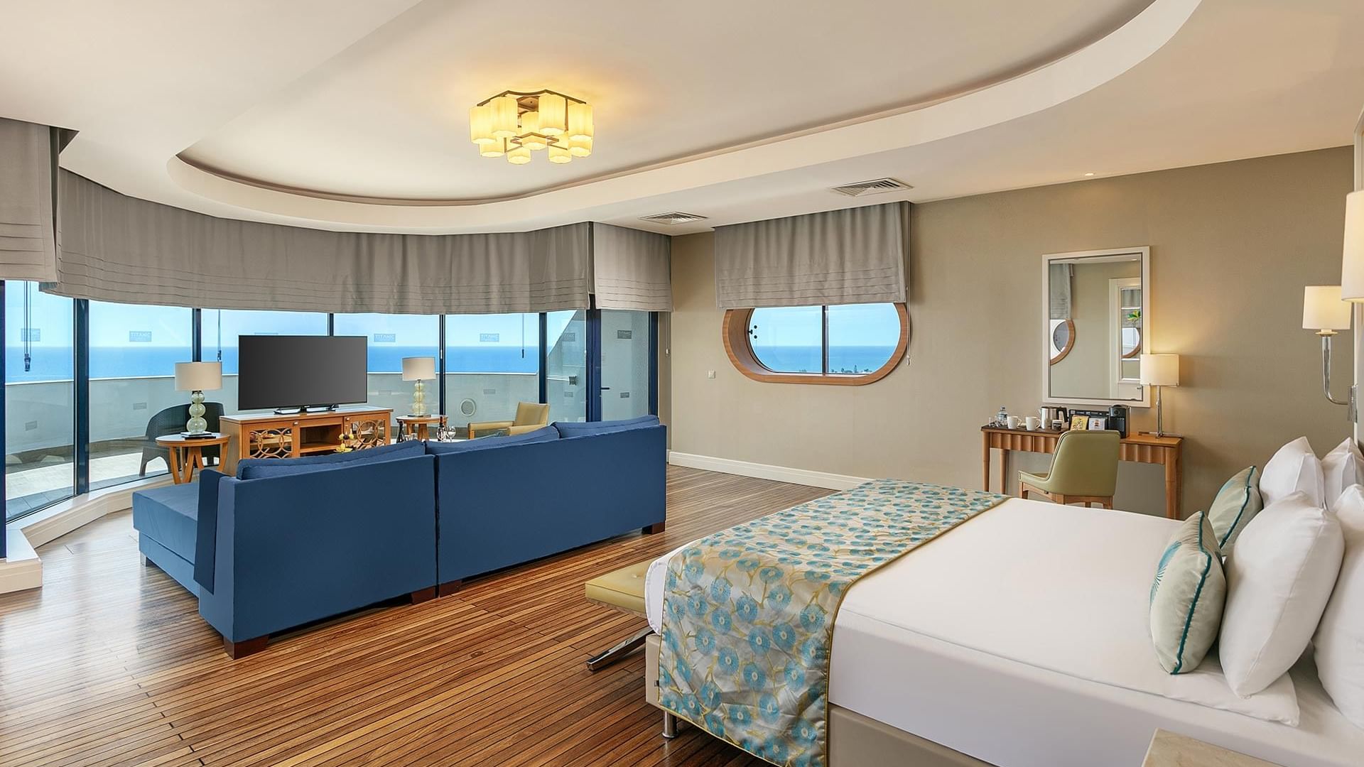 Titanic Deluxe Lara – Admiral Suite’te deniz manzaralı, mavi kanepeli ve beyaz yataklı oda.