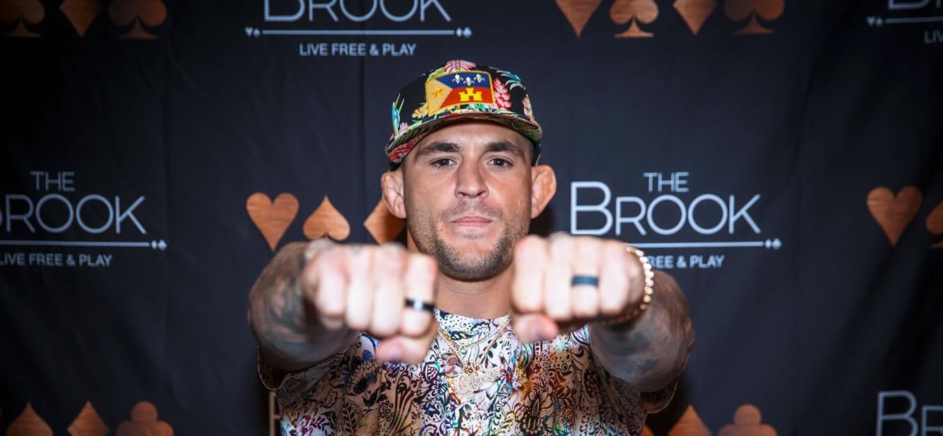 UFC fighter Dustin Poirier raising both fists toward camera in front of branded backdrop wall at The Brook