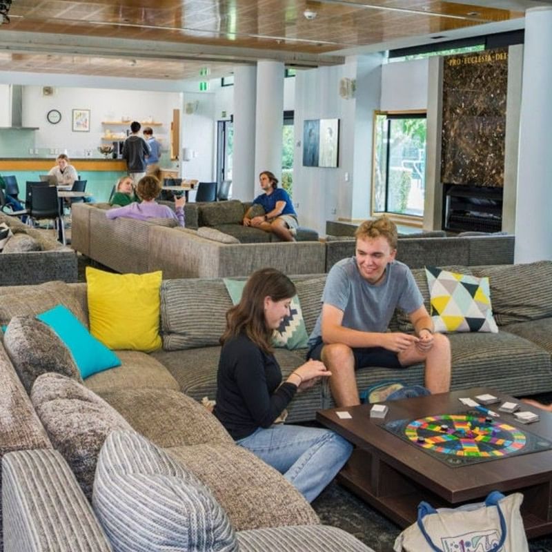 Guests socializing and playing board games in a spacious communal lounge with colorful cushions and wooden ceiling beams.