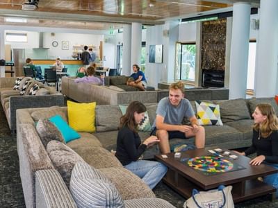 Guests socializing and playing board games in a spacious communal lounge with colorful cushions and wooden ceiling beams.