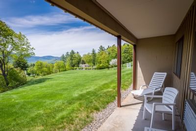 Fox Ridge Conway NH room with patio with white chairs, a grassy hill, trees, and mountains under a partly cloudy sky.