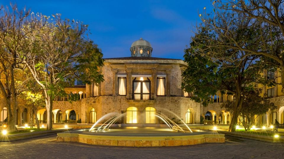 Illuminated stone facade and central fountain at dusk, featuring elegant arches at Quinta Real Aguascalientes