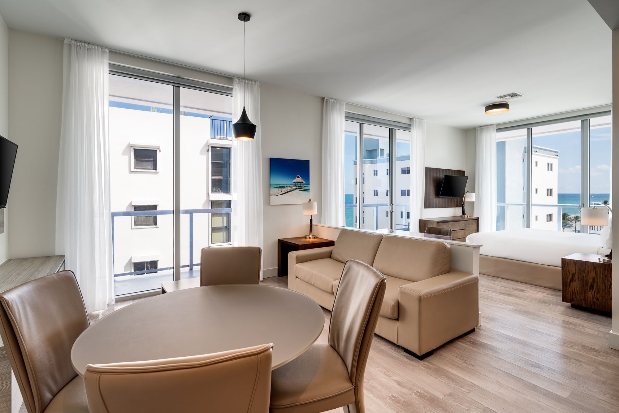 A wide-angle view of a hotel suite with a dining area, sofa, and large windows overlooking the ocean.