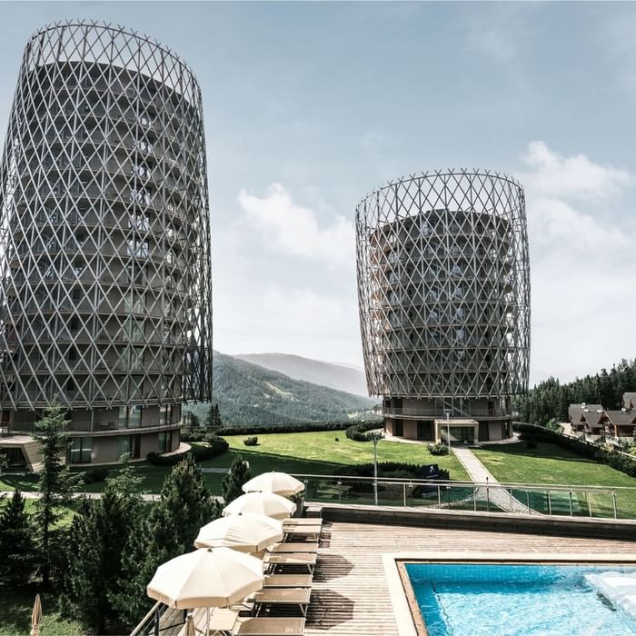 Modern towers in a green terrain with a pool and lounge chairs under a sky at Falkensteiner Hotels