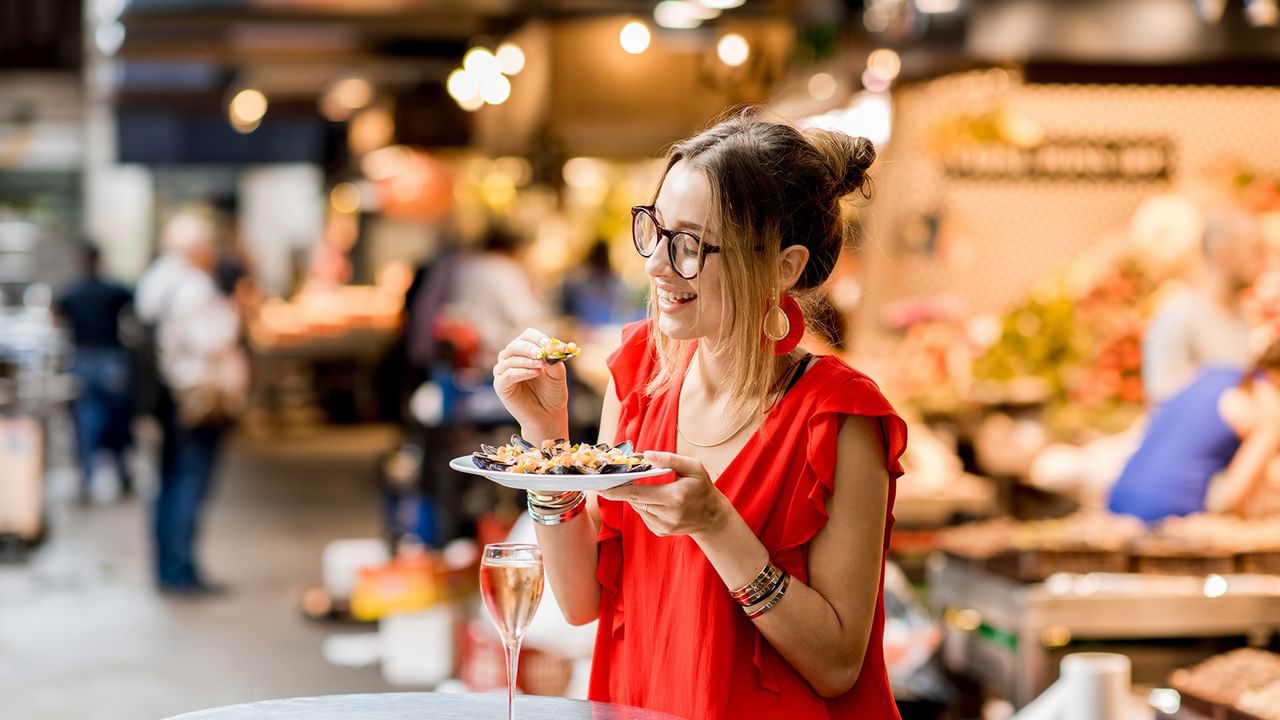 Woman in red dress and glasses eating food at a market with a glass of wine.