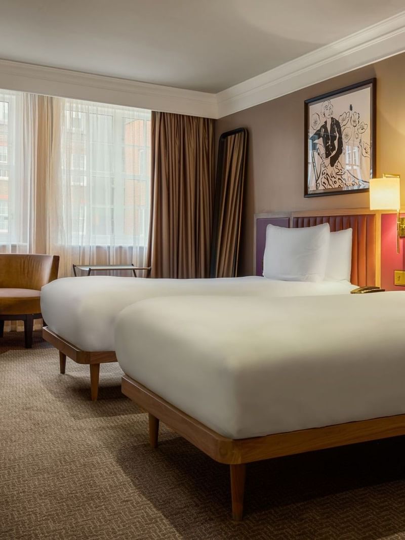 A well-furnished hotel room with two beds, lamps, and a window in London.