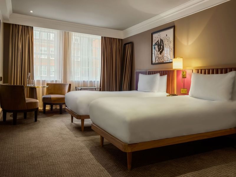 A well-furnished hotel room with two beds, lamps, and a window in London.