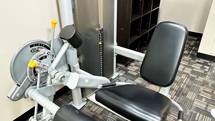 A weight training machine with a black seat and gray frame in a fitness center with cubby storage and carpeted flooring.