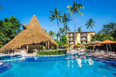 Resort with blue pool, thatched roof bar, lounge chairs, umbrellas, and palm trees under a clear blue sky.