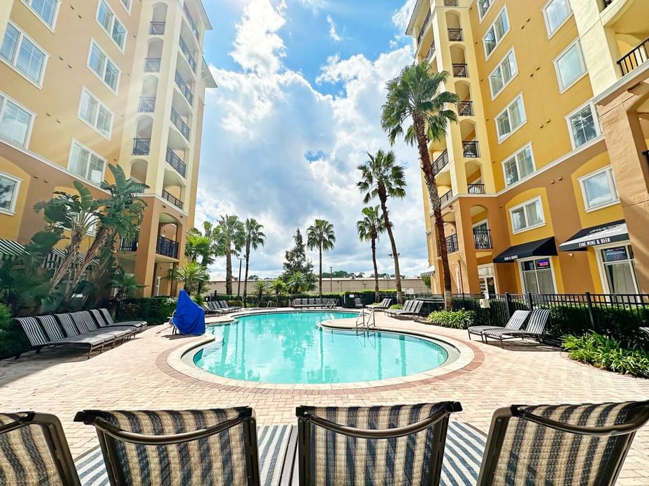 A pool surrounded by lounge chairs and palm trees, with a building in the background.