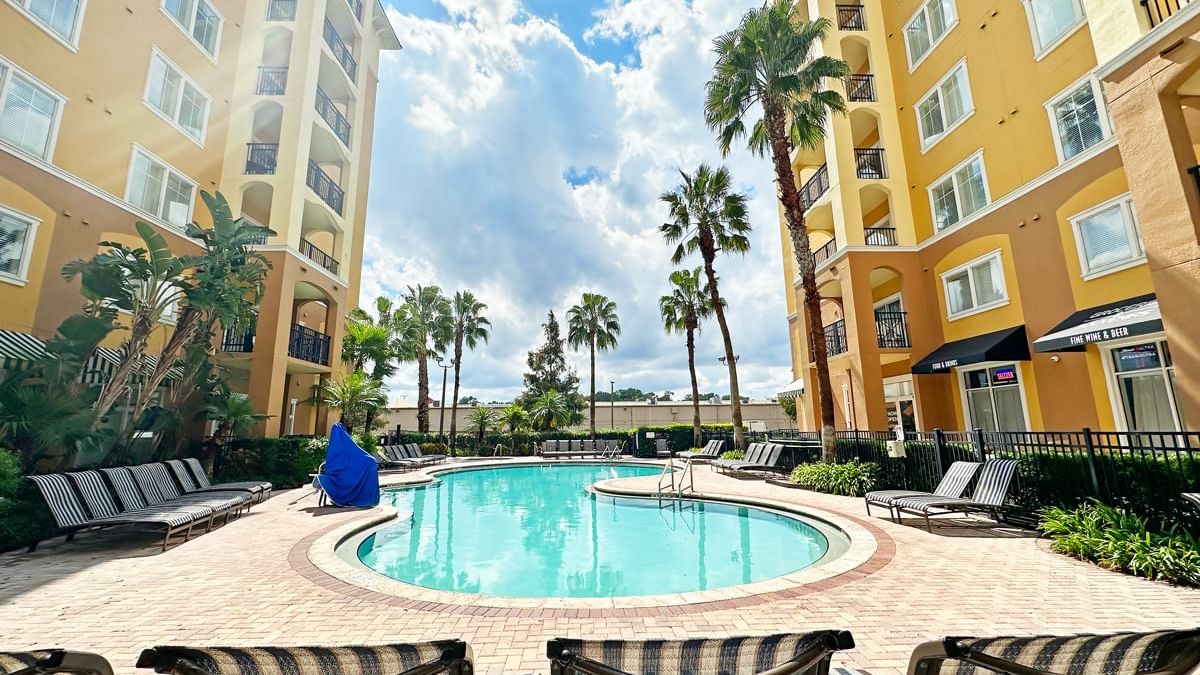 A pool area with a round pool, lounge chairs, and palm trees, surrounded by a yellow multi-story building.